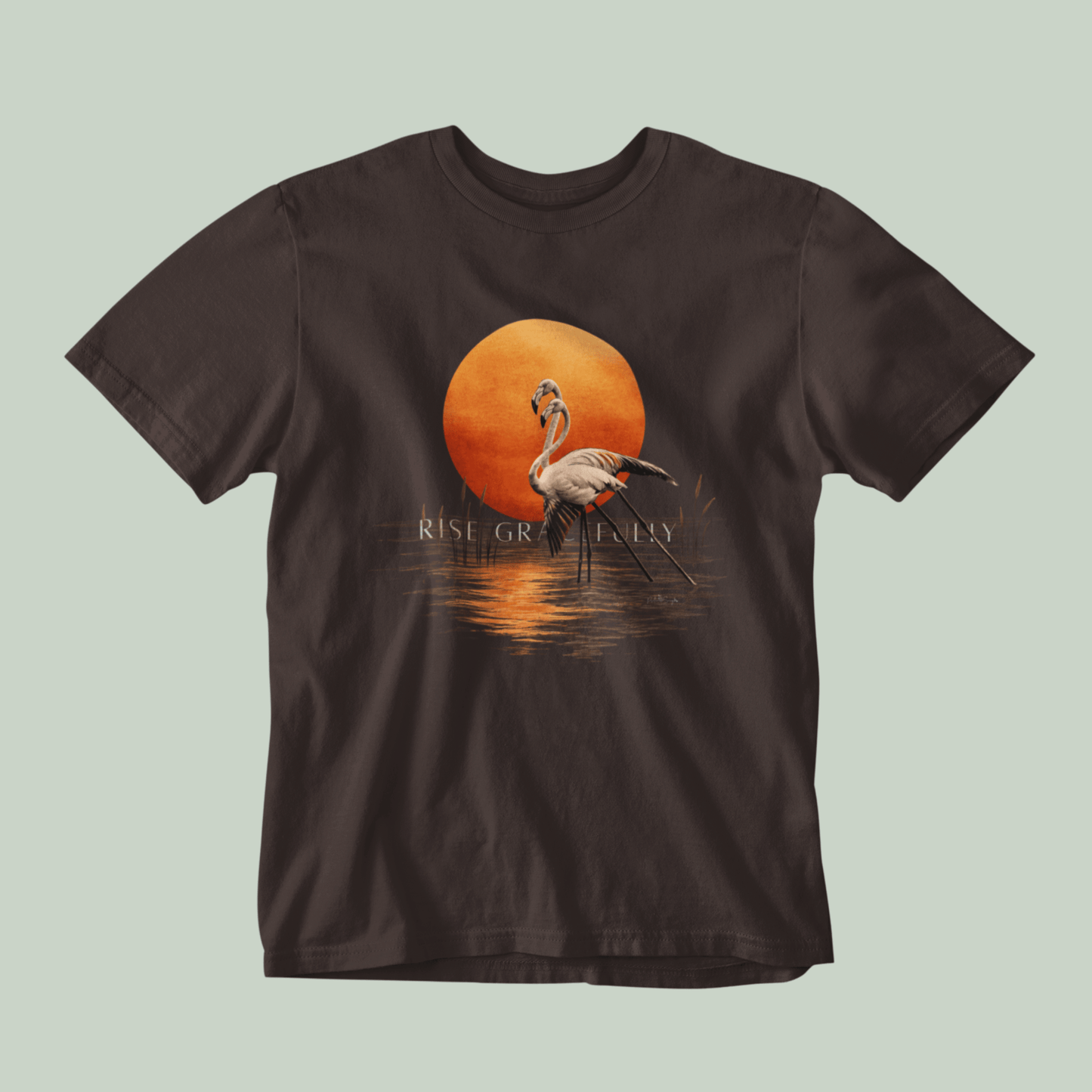 A brown Tucktribe short-sleeve crewneck t-shirt featuring a graphic print of a pink flamingo standing in front of a large orange sun over water, with the text 'RISE GRACEFULLY' printed below the sun.