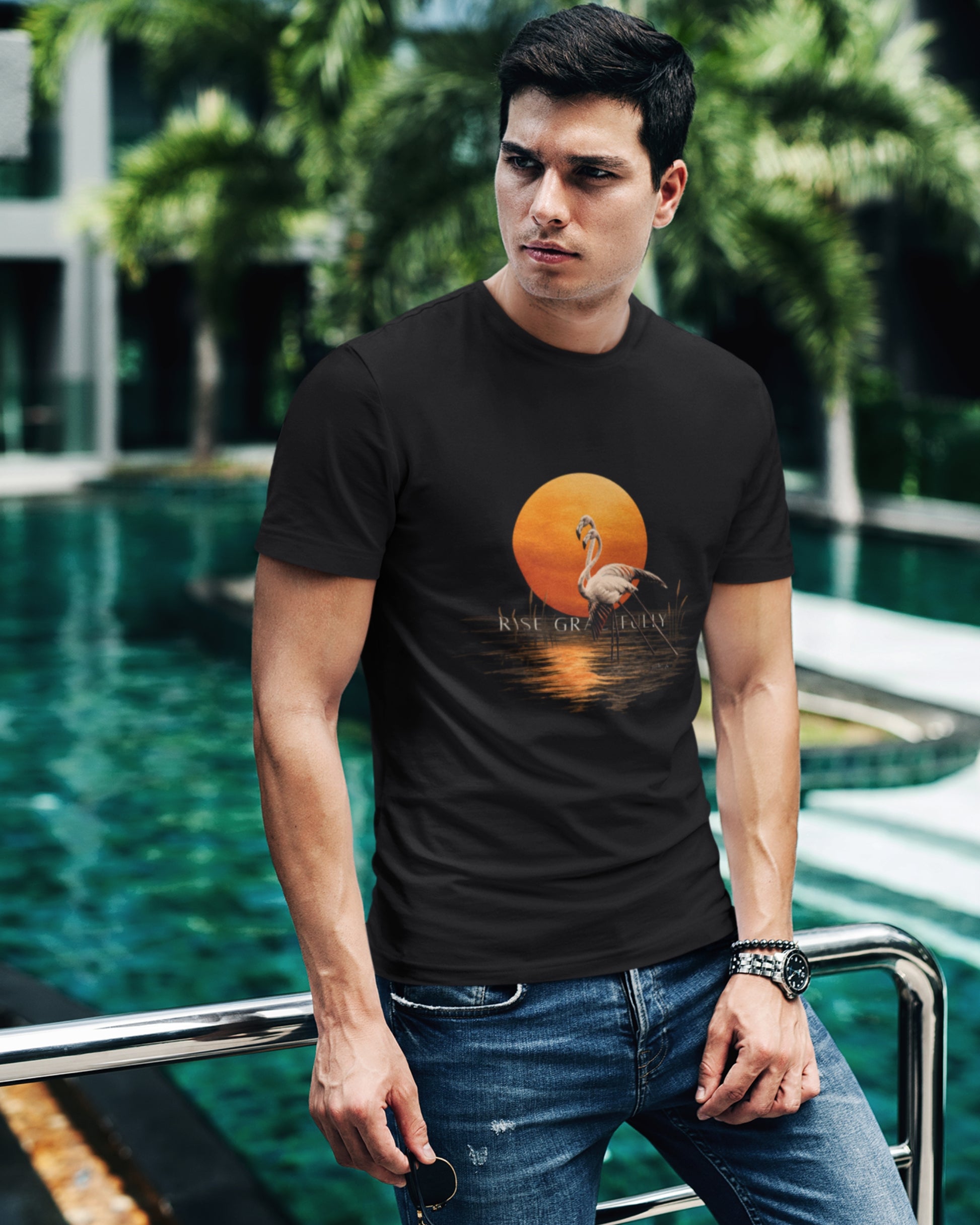 A man wearing a black tucktribe graphic t-shirt with a flamingo and orange sun design, standing by a swimming pool with palm trees in the background.