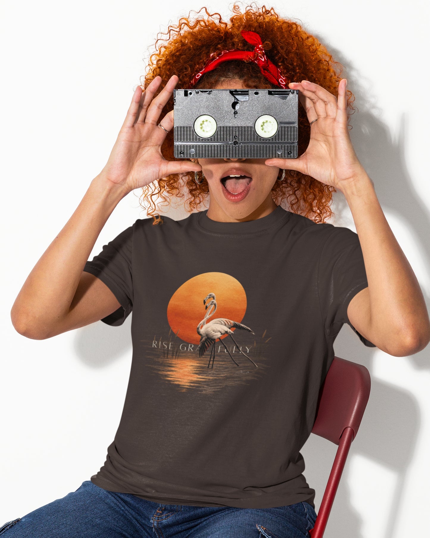 A person with curly red hair holds a VHS tape over their eyes like a camera, wearing a dark brown tucktribe graphic t-shirt with a flamingo sunset print and blue jeans, seated on a red chair against a white background.