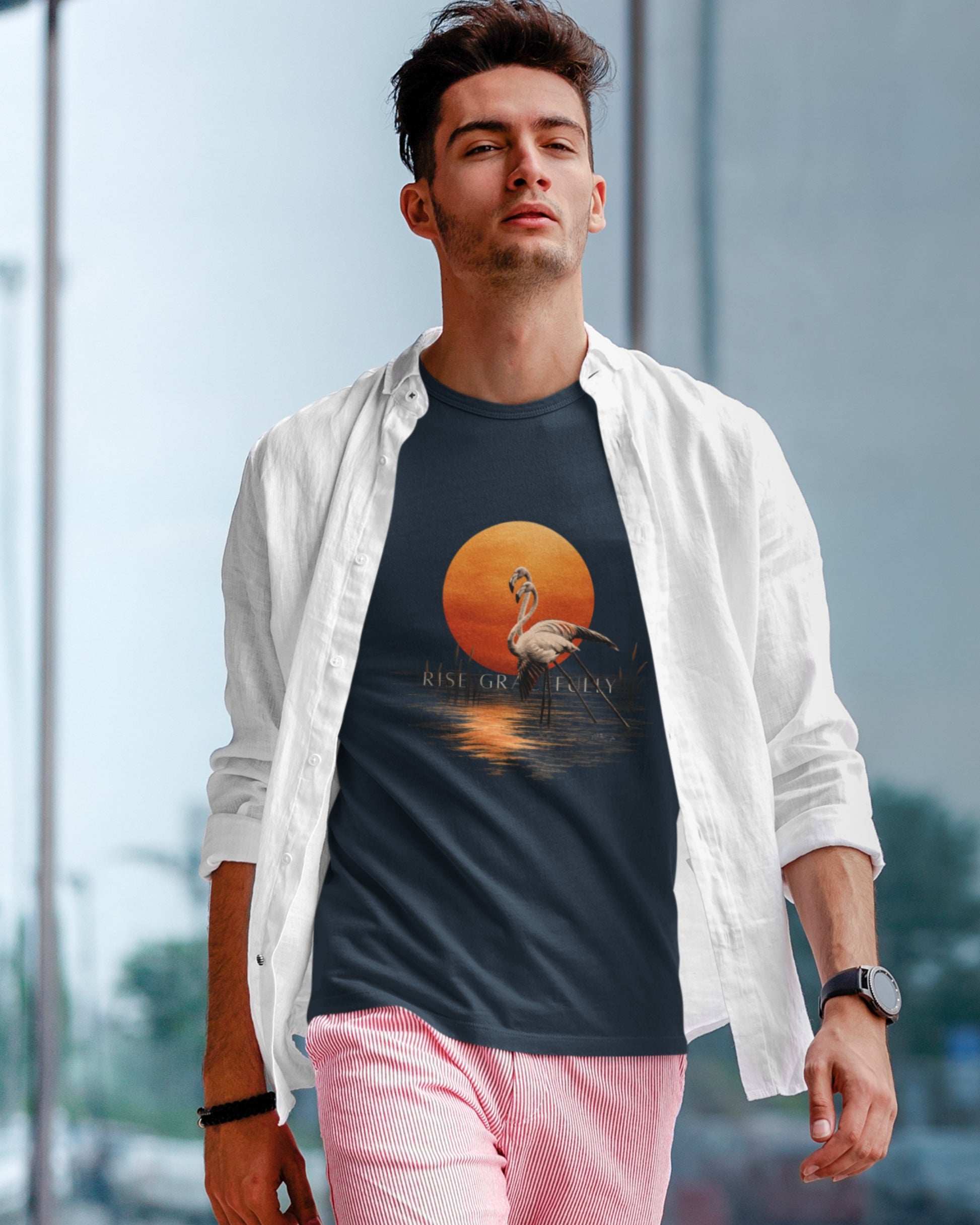 A man wearing a Tucktribe navy blue graphic t-shirt with a flamingo and sunset print, styled with an open white button-down shirt and pink pants, standing in front of a blurred modern glass building background.