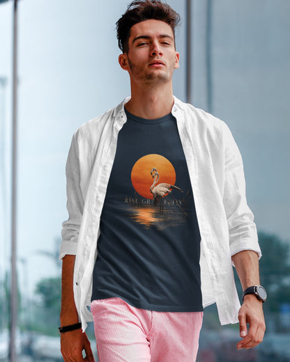 A man wearing a Tucktribe navy blue graphic t-shirt with a flamingo and sunset print, styled with an open white button-down shirt and pink pants, standing in front of a blurred modern glass building background.