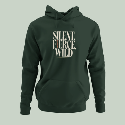 A dark forest green pullover hoodie with a front kangaroo pocket and drawstring hood, featuring the text 'SILENT, FIERCE, WILD' printed in a cream-colored stencil font on the chest. The hoodie is displayed on a solid light sage green background.
