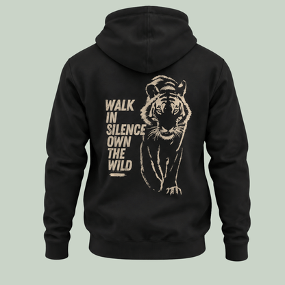 A black pullover hoodie shown from the back, featuring a large distressed beige graphic of a walking tiger and the text 'WALK IN SILENCE OWN THE WILD' on a light sage green background.