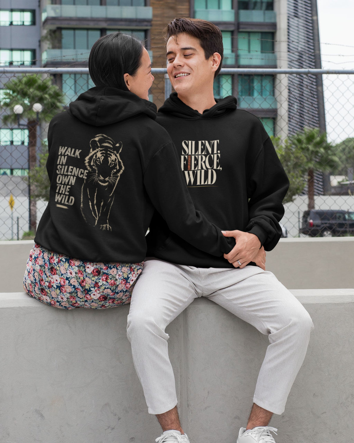 Two people sitting on a concrete ledge outdoors, both wearing matching black hoodies with graphic prints. The person on the left shows the back of the hoodie with a tiger illustration and the text 'WALK IN SILENCE OWN THE WILD.