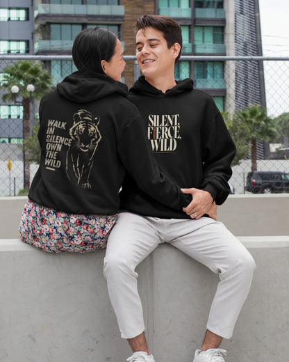 Two people sitting on a concrete ledge outdoors, both wearing matching black hoodies with graphic prints. The person on the left shows the back of the hoodie with a tiger illustration and the text 'WALK IN SILENCE OWN THE WILD.