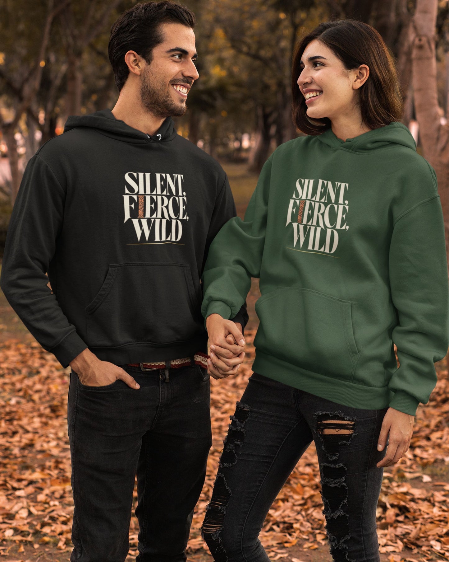 A man and a woman standing outdoors in a park with fallen leaves, wearing matching hoodies with the text 'SILENT. FIERCE, WILD.