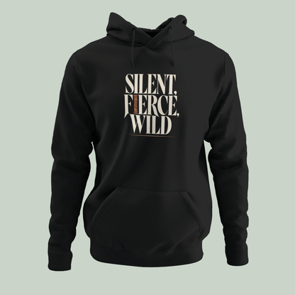 A black pullover hoodie with a front kangaroo pocket and drawstring hood, featuring the text 'SILENT, FIERCE, WILD' printed in a distressed cream and brown font on the chest, set against a solid pale sage green background.