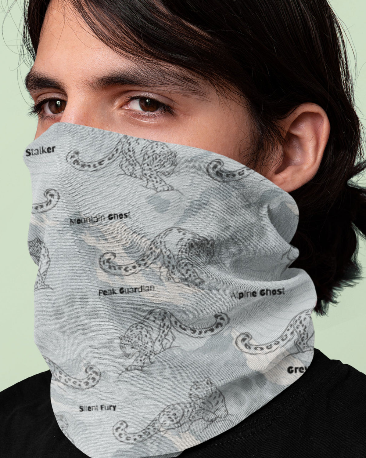 A close-up of a person wearing a light gray neck gaiter mask with a black and white snow leopard pattern and text labels.