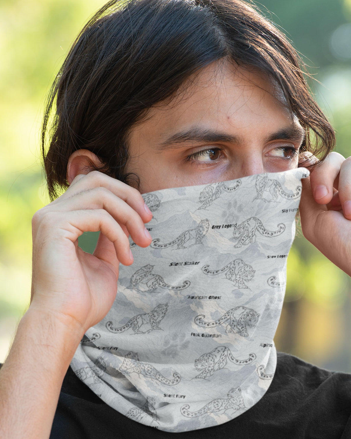 A close-up of a person outdoors wearing a light gray patterned neck gaiter or face covering, holding it in place with both hands. The fabric features a repeating gray and black abstract print of snowleopards on a light gray background.