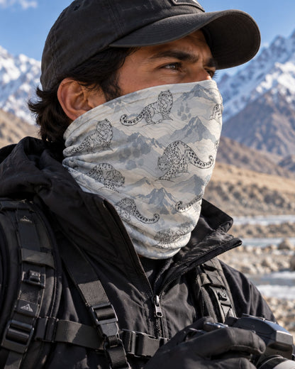 A close-up of a man outdoors wearing a light gray and white patterned neck gaiter with a black and white mountain and snow leopard print, along with a black cap and jacket, with snow-capped mountains in the background.