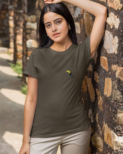 Woman wearing a olive green tucktribe t-shirt with a toucan bird embroidery, standing against a stone wall.