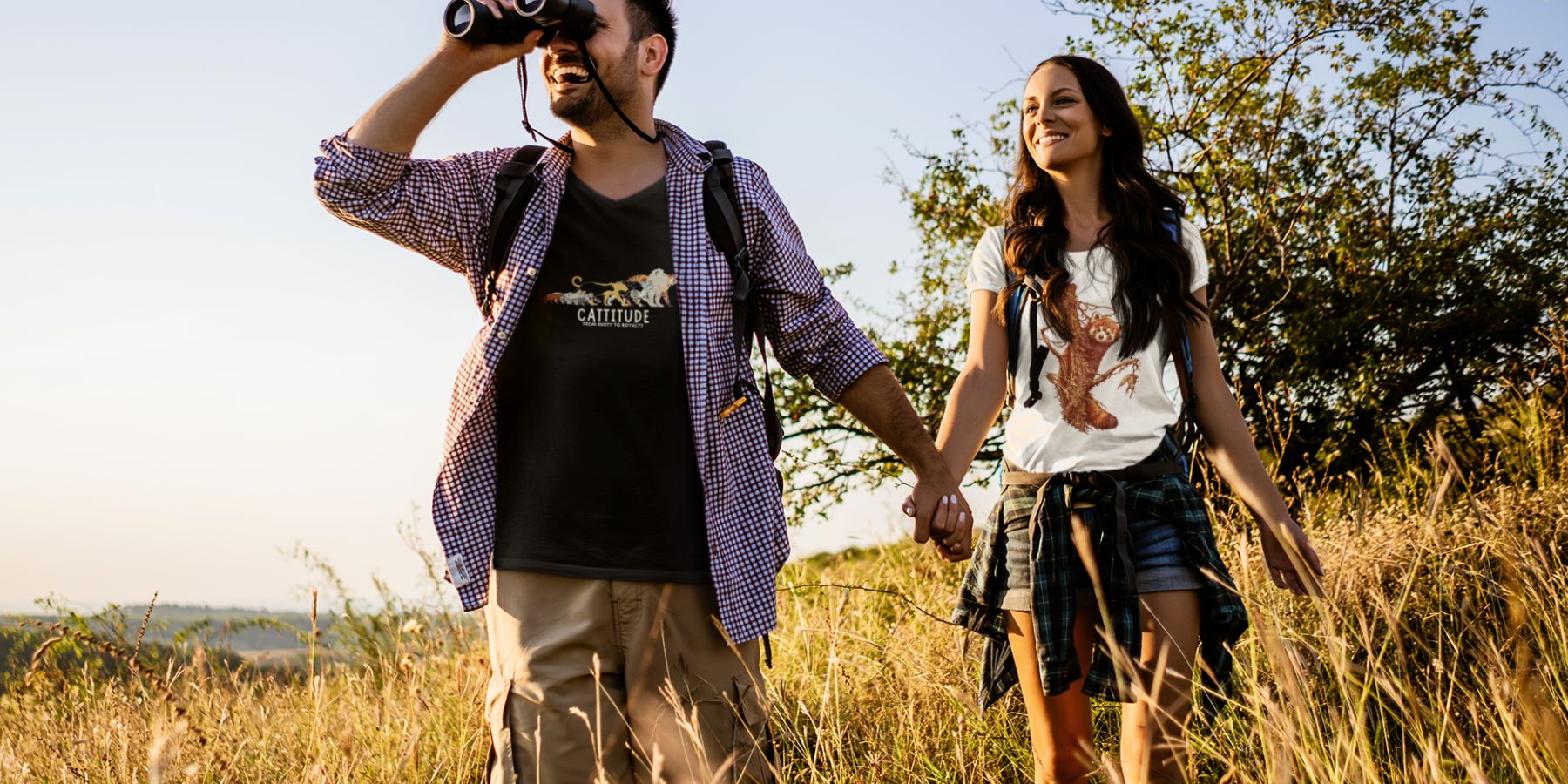 Two people wearing tucktribe tshirts walking through a field with binoculars and a camera, enjoying nature.