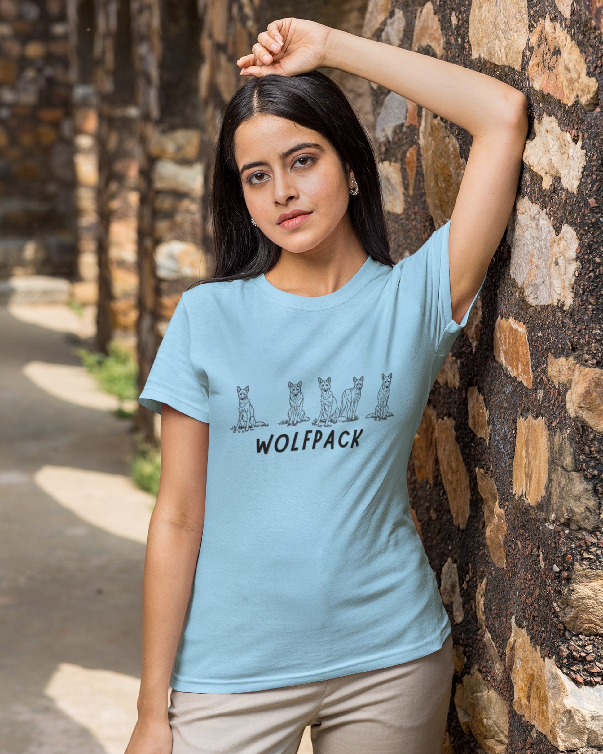 Woman wearing a light blue t-shirt with 'Wolfpack' design, standing against a stone wall.