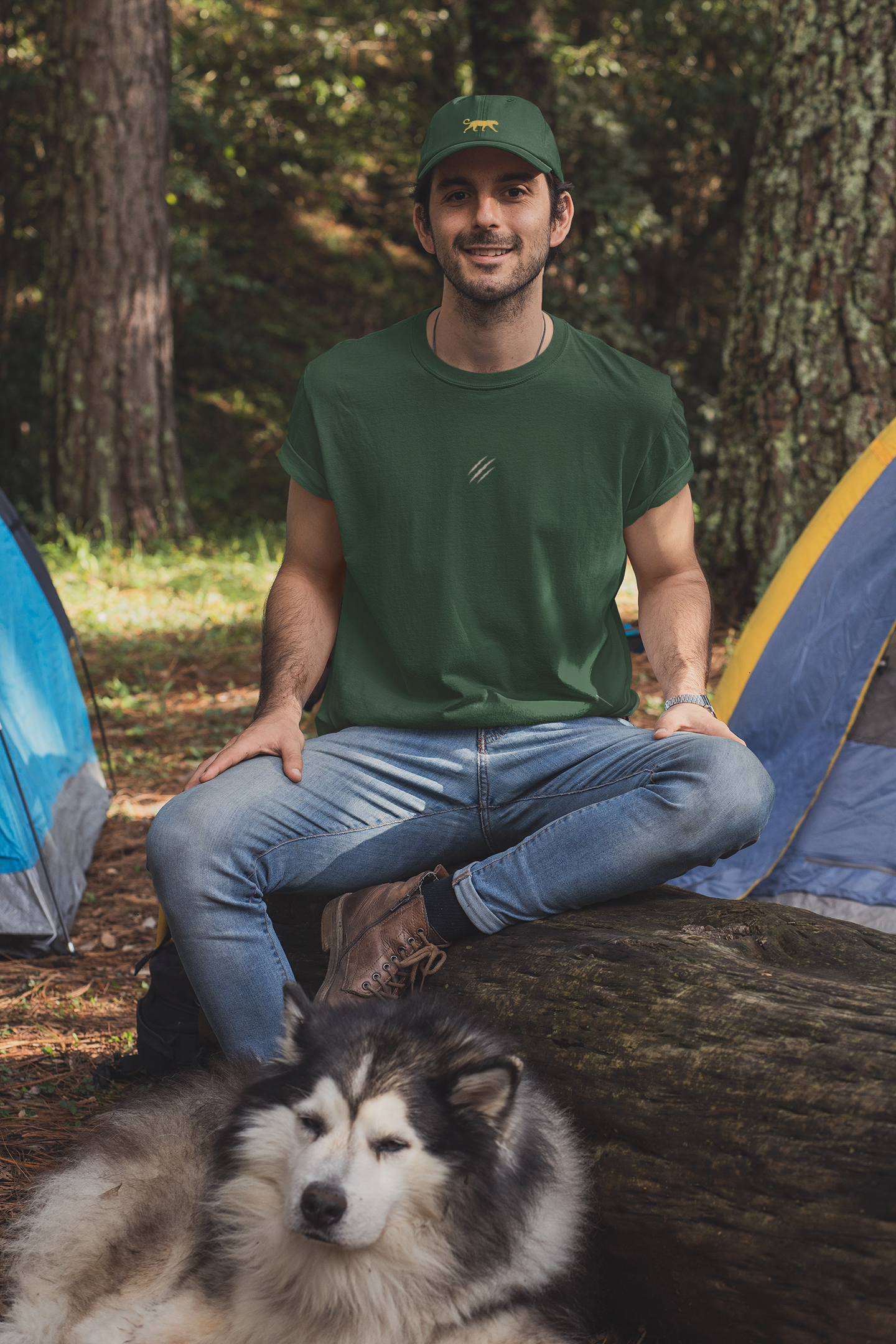 Man wearing a tucktribe bottle green t-shirt and cap sitting with a dog in a forest setting near tents.