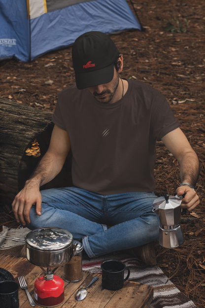 Man wearing a tucktribe brown t-shirt with claw scratch embroidery camping, sitting by a campfire with a coffee percolator and food items.