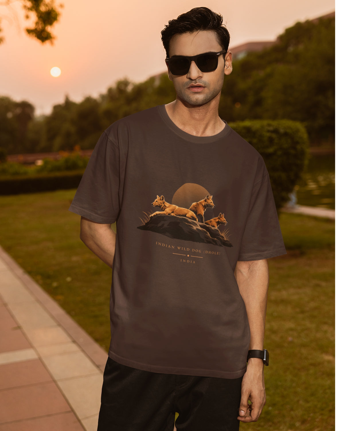 Man wearing a brown Dhole wildlife T-shirt featuring three Indian wild dogs on a rocky landscape with sunset-inspired artwork