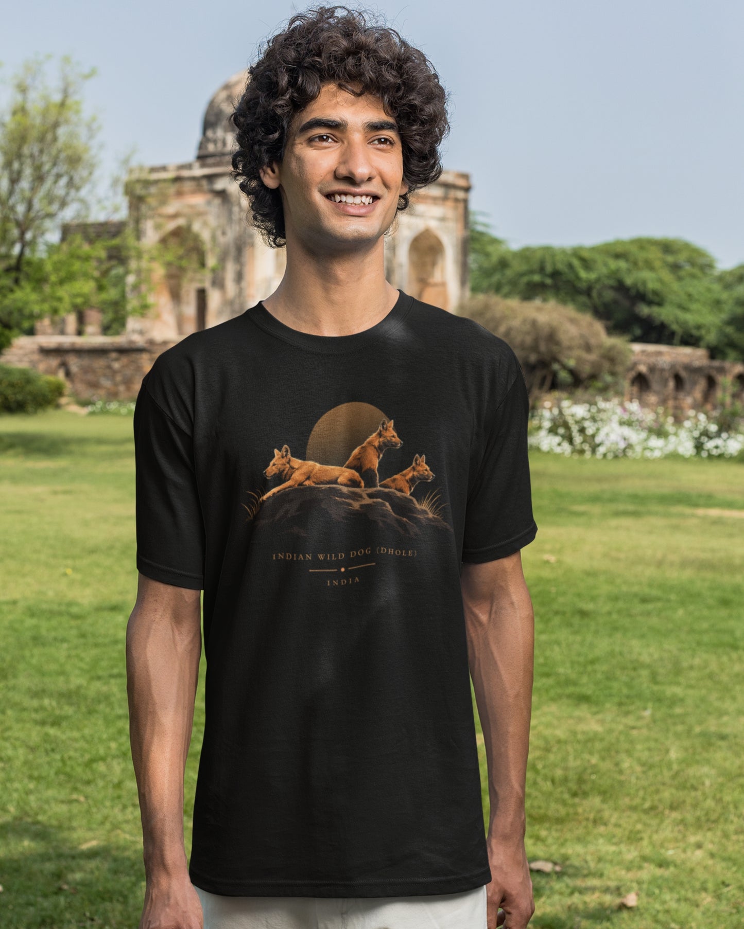 Man wearing a black Dhole wildlife T-shirt featuring three Indian wild dogs on a rocky landscape with sunset-inspired artwork