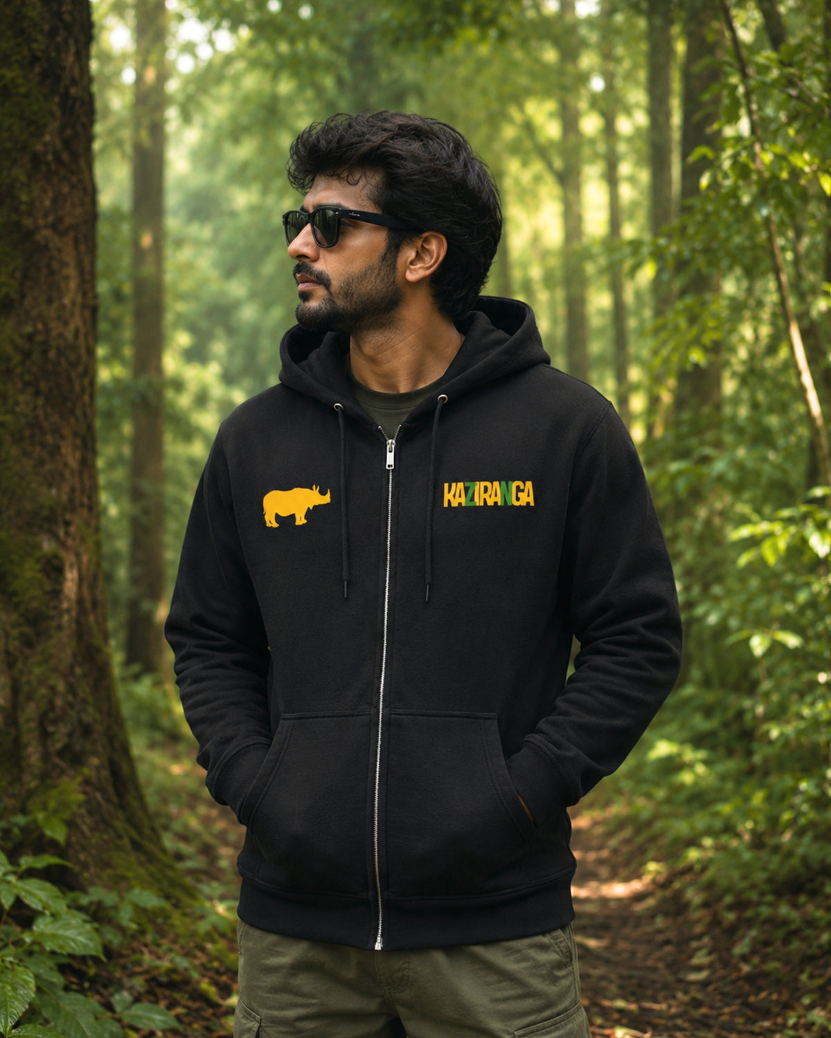 A man wearing a black tucktribe zip-up hoodie with yellow graphics stands in a forest. The hoodie features a yellow rhinoceros silhouette on the left chest and the word 'KAZIRANGA' in yellow on the right chest.