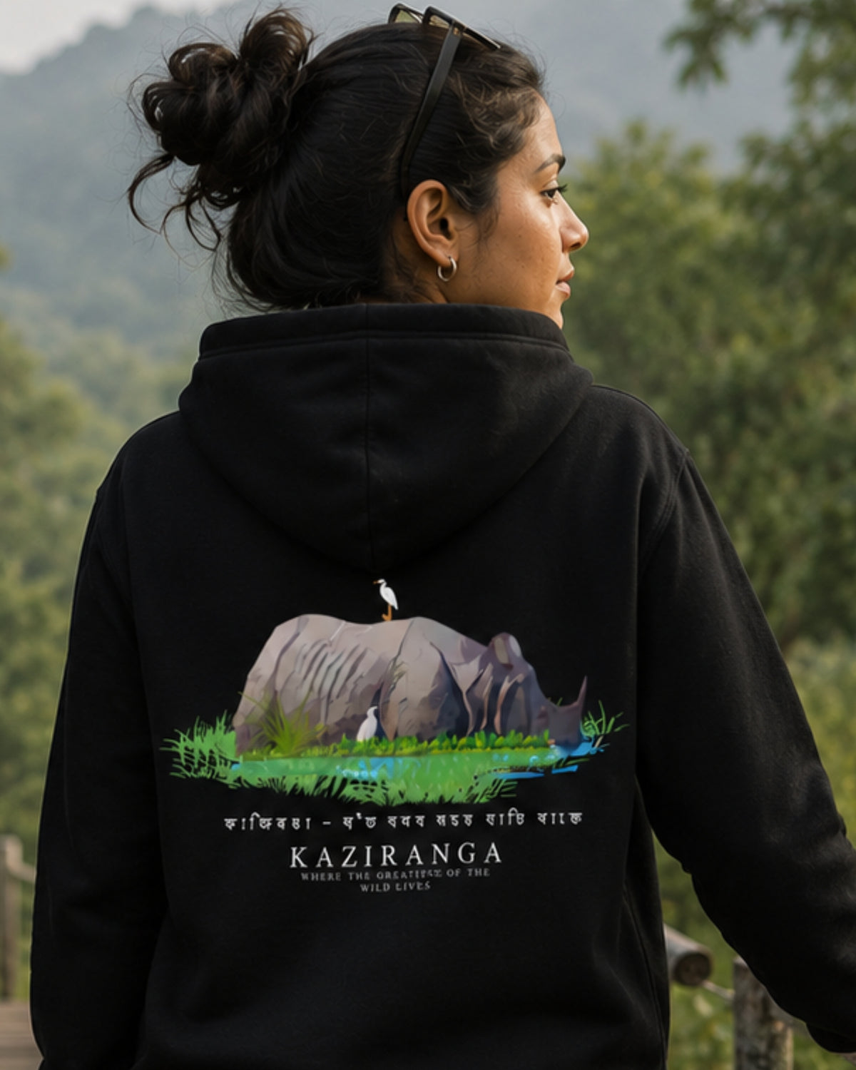 A woman wearing a black tucktribe hoodie with a large back graphic of a rhinoceros and bird, photographed outdoors against a blurred green mountain background.