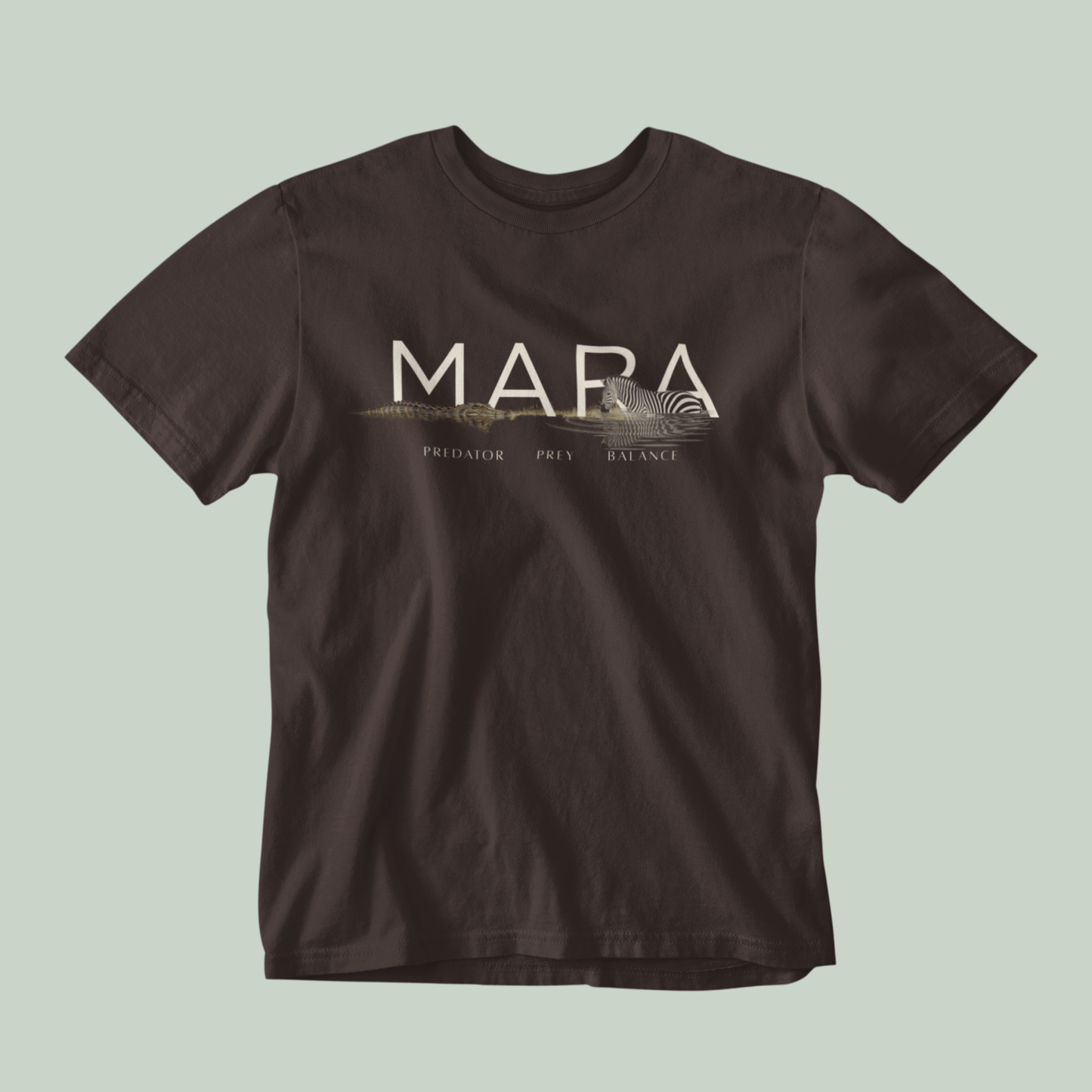 A brown Tucktribe short-sleeve crewneck t-shirt with a beige graphic print on the chest featuring the word 'MARA' and smaller text 'PREDATOR - PREY- BALANCE' with a Zebra and crocodile illustration, displayed against a solid light sage green background.
