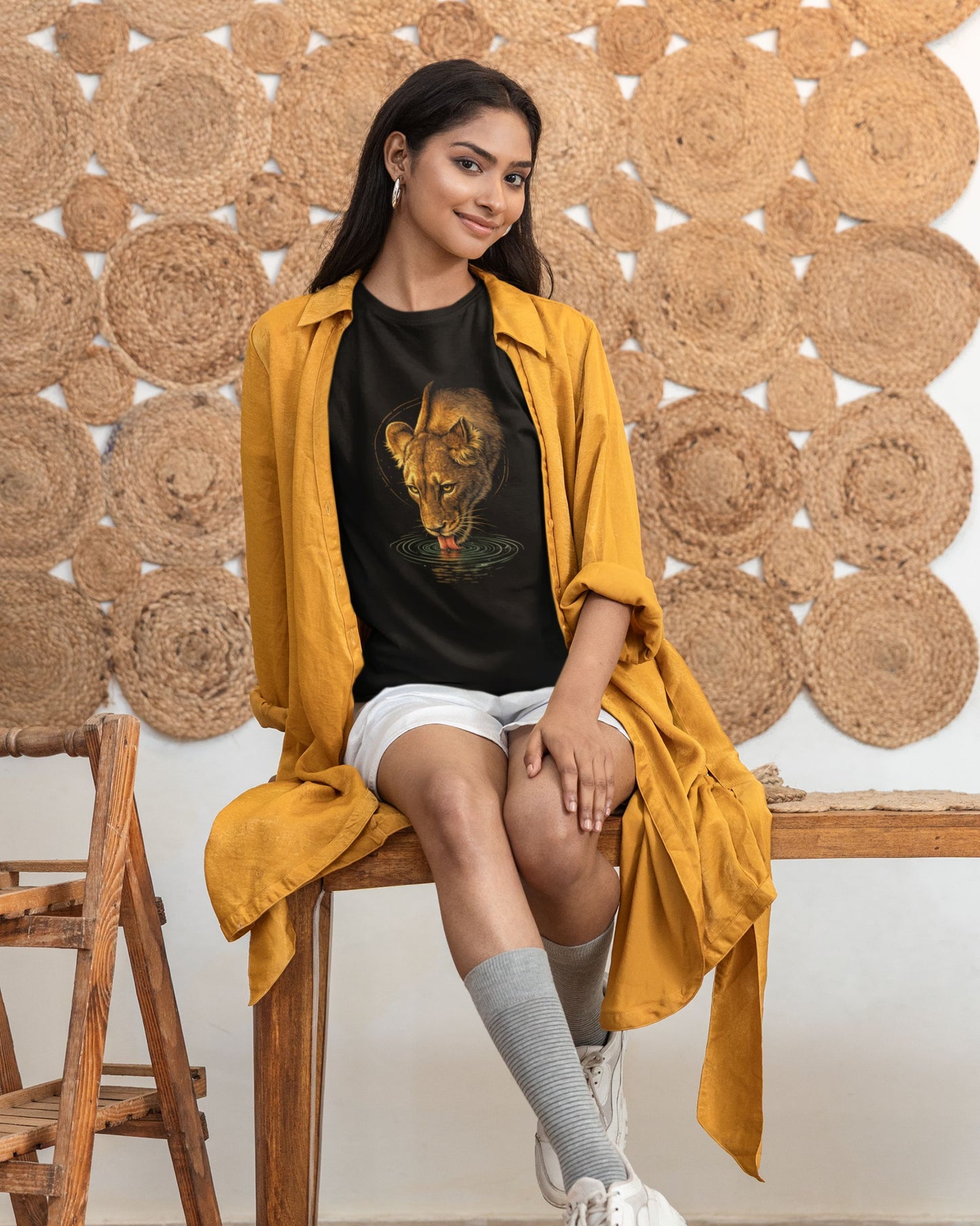 Woman wearing a black tucktribe t-shirt with a lion graphic and a mustard yellow cardigan sitting on a wooden chair.