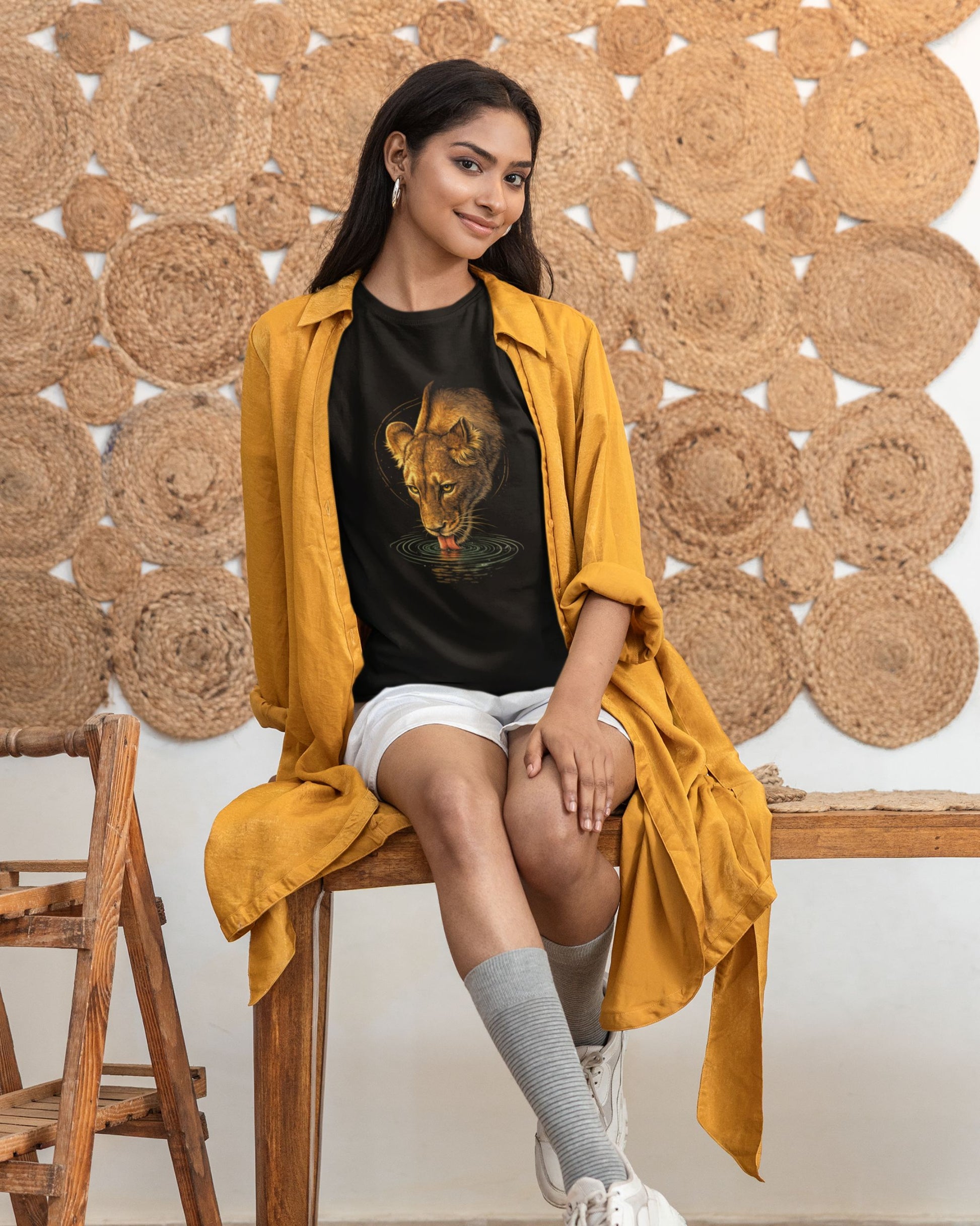 Woman wearing a black tucktribe t-shirt with a lion graphic and a mustard yellow cardigan sitting on a wooden chair.