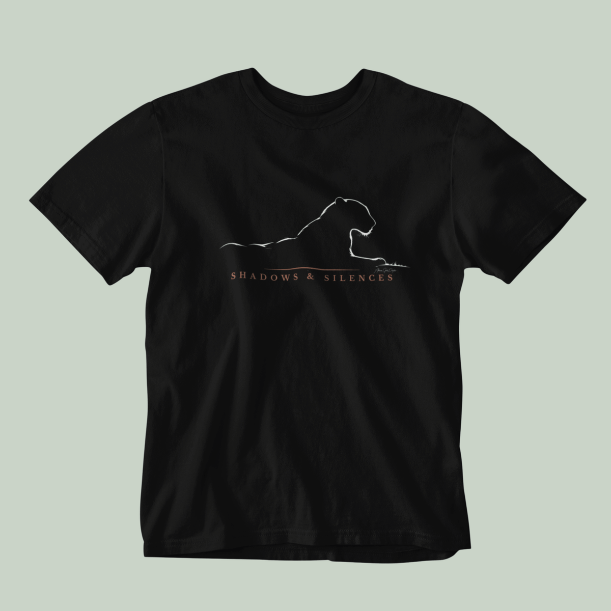 A black short-sleeve crewneck t-shirt featuring a white line art graphic of a leopard from jawai and the text 'SHADOWS & SILENCES' printed on the chest, displayed against a solid light sage green background.