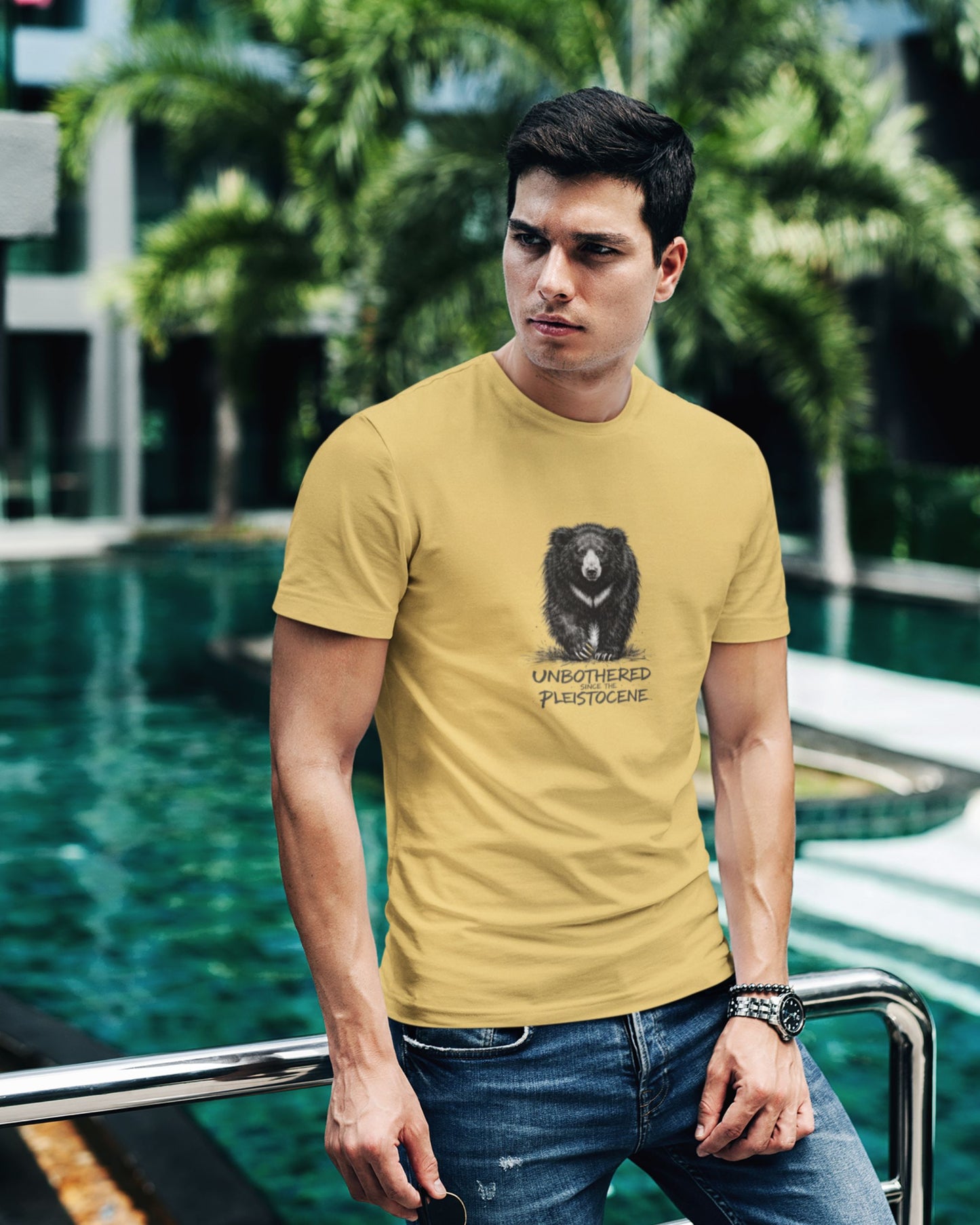 Man wearing a beige tucktribe t-shirt with a sloth bear graphic design and text, standing by a pool with greenery in the background.