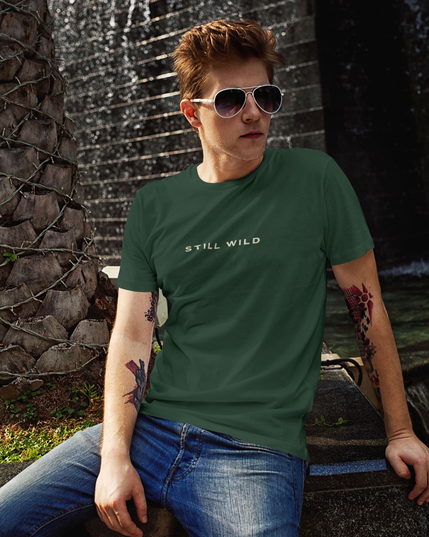 Person wearing a bottle green tucktribe t-shirt with 'STILL WILD' text, sitting outdoors.