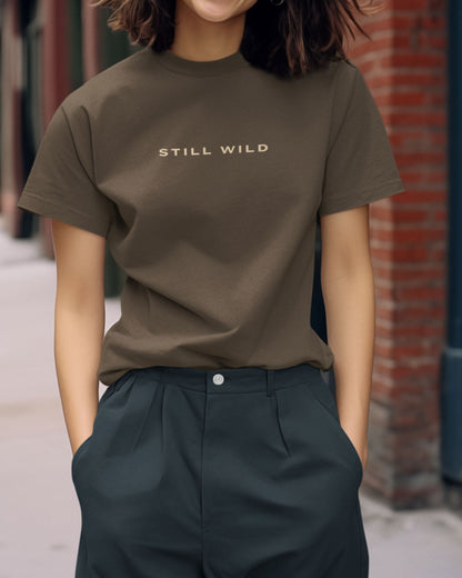 Person wearing a olive green tucktribe t-shirt with 'STILL WILD' text, standing against a brick wall.