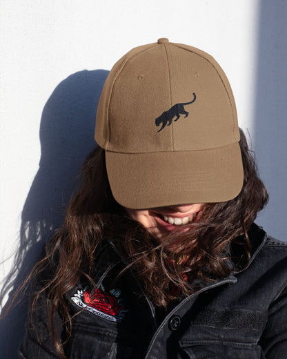 Person wearing a  brown tucktribe cap with a black panther logo against a white wall.