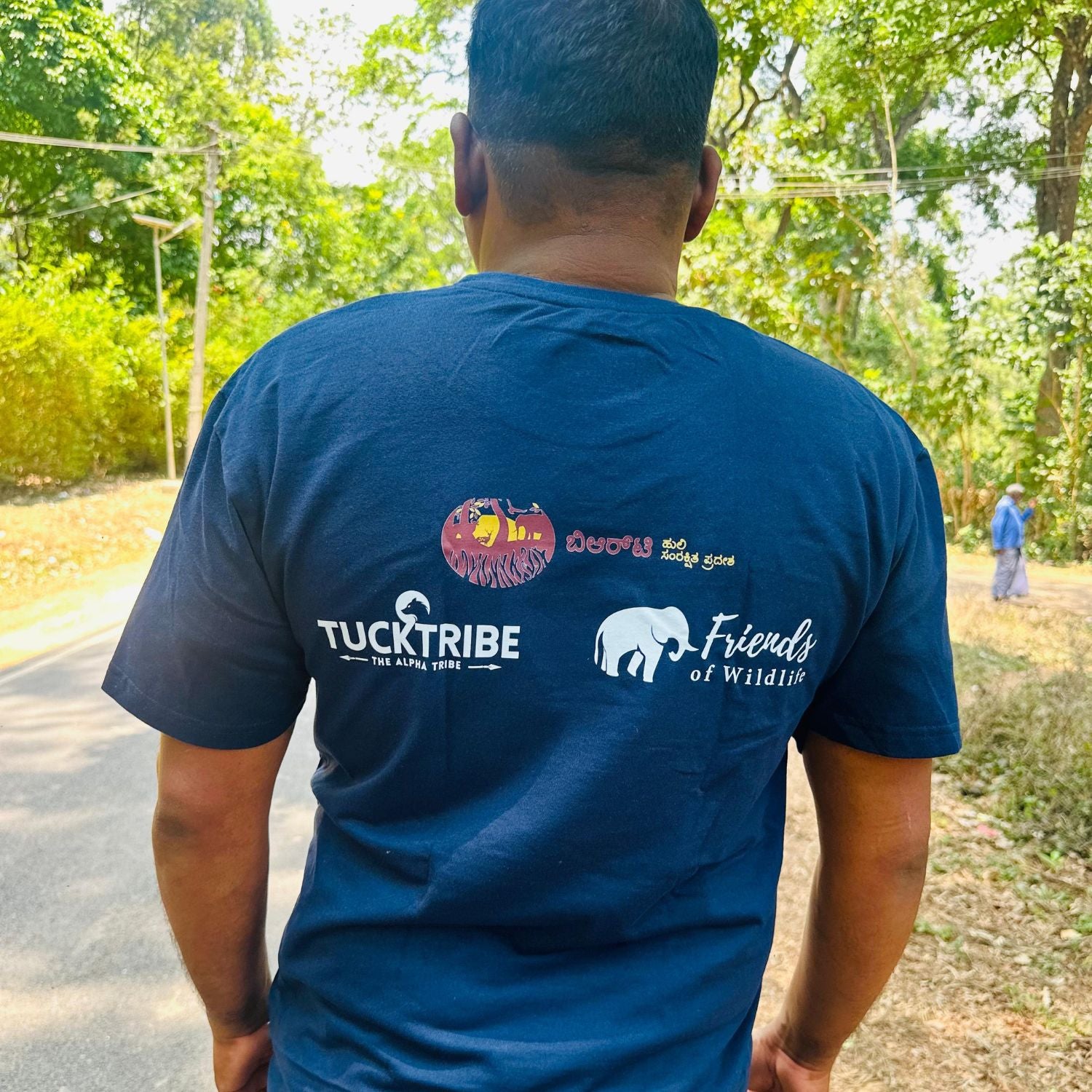 Person wearing a blue tucktribe t-shirt with logos on the back, standing outdoors with trees in the background.