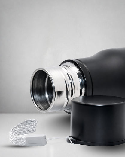 closeup of a matte black insulated tucktribe water bottle with a matching black screw cap opend to and a white wrist strap, featuring a engraving of a cobra and the text 'BEYOND FEAR' on the front, set against a warm interior.