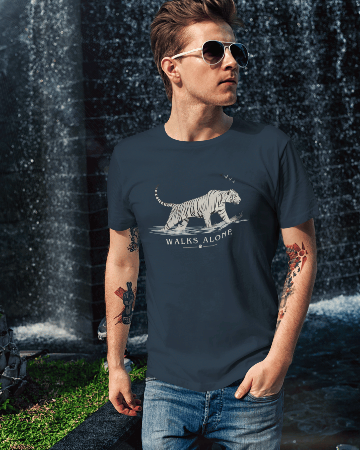 A man wearing a navy blue t-shirt with a white tiger graphic and the text 'WALKS ALONE' stands in front of a waterfall. The t-shirt features a detailed white tiger illustration and the phrase 'WALKS ALONE' printed below it.