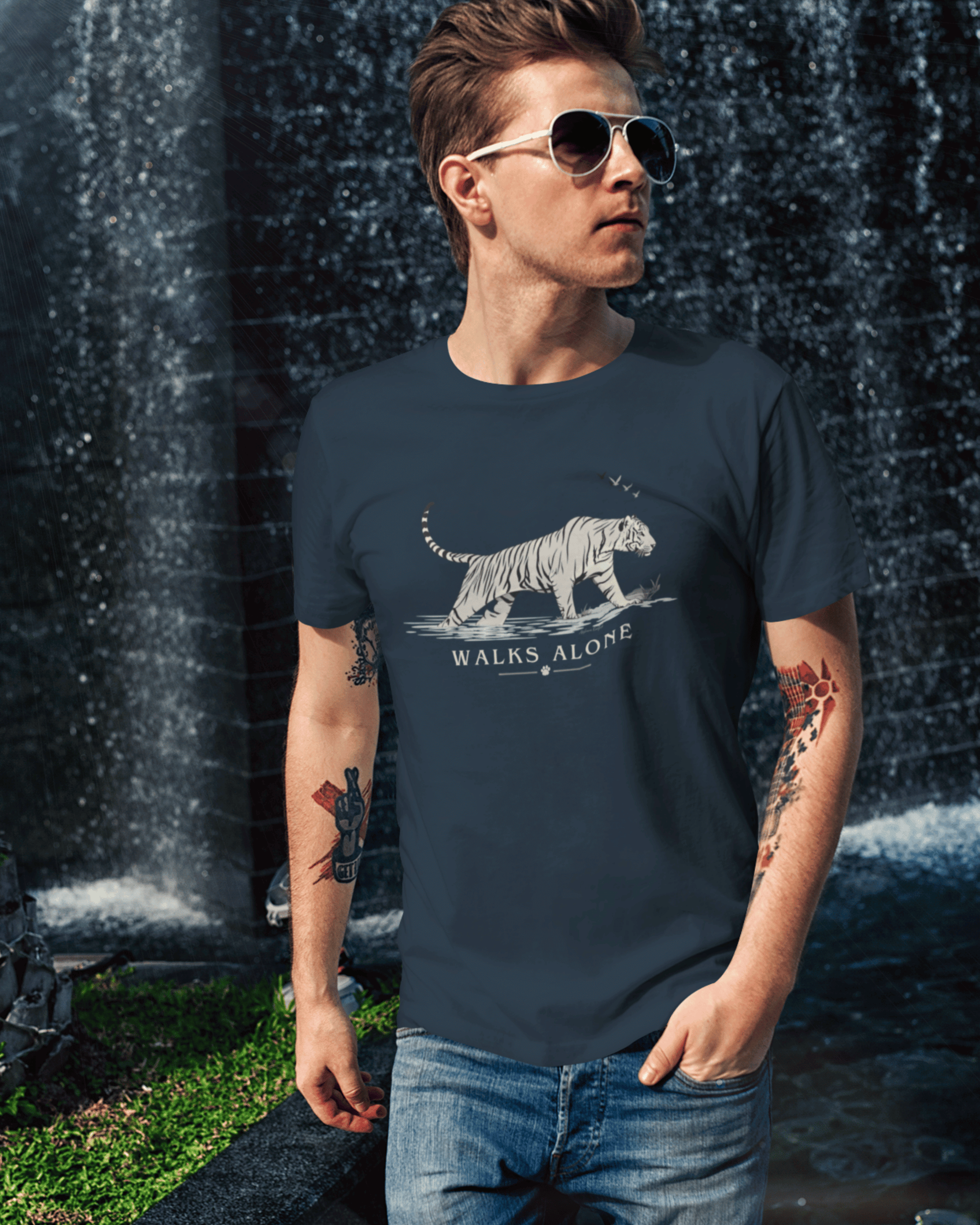 A man wearing a navy blue t-shirt with a white tiger graphic and the text 'WALKS ALONE' stands in front of a waterfall. The t-shirt features a detailed white tiger illustration and the phrase 'WALKS ALONE' printed below it.