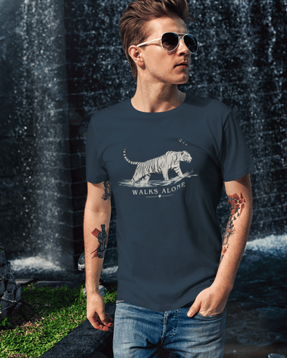 A man wearing a navy blue t-shirt with a white tiger graphic and the text 'WALKS ALONE' stands in front of a waterfall. The t-shirt features a detailed white tiger illustration and the phrase 'WALKS ALONE' printed below it.