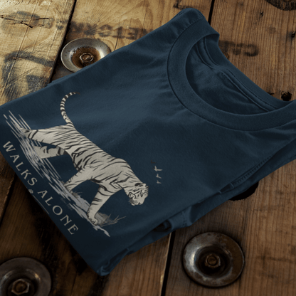 A navy blue folded t-shirt with a white graphic of a tiger and the text 'WALKS ALONE', displayed on a rustic wooden background with metal rivets.