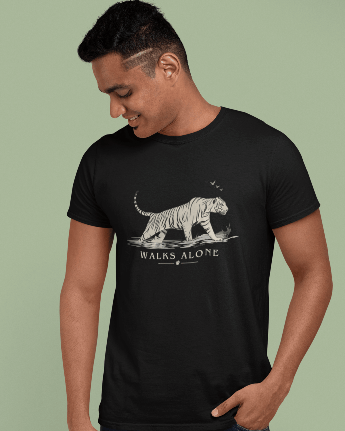 A man wearing a black short-sleeve t-shirt with a white graphic of a walking tiger and the text 'WALKS ALONE' on the front, set against a solid sage green background.