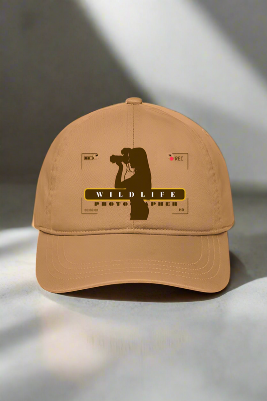 The TuckTribe WILDLIFE PHOTOGRAPHER(F) PRINTED BASEBALL CAP, a beige cap for women, showcases a silhouette of a wildlife photographer with a camera and the bold WILDLIFE PHOTOGRAPHY surrounded by recording icons, stylishly resting on a gray surface.